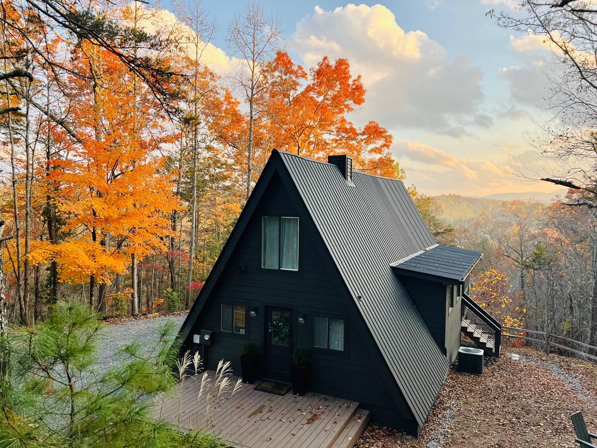 The Little A at golden hour: a dark A-frame with a standing-seam roof, a wood deck out front, orange fall maples on the left and a hazy mountain ridge behind